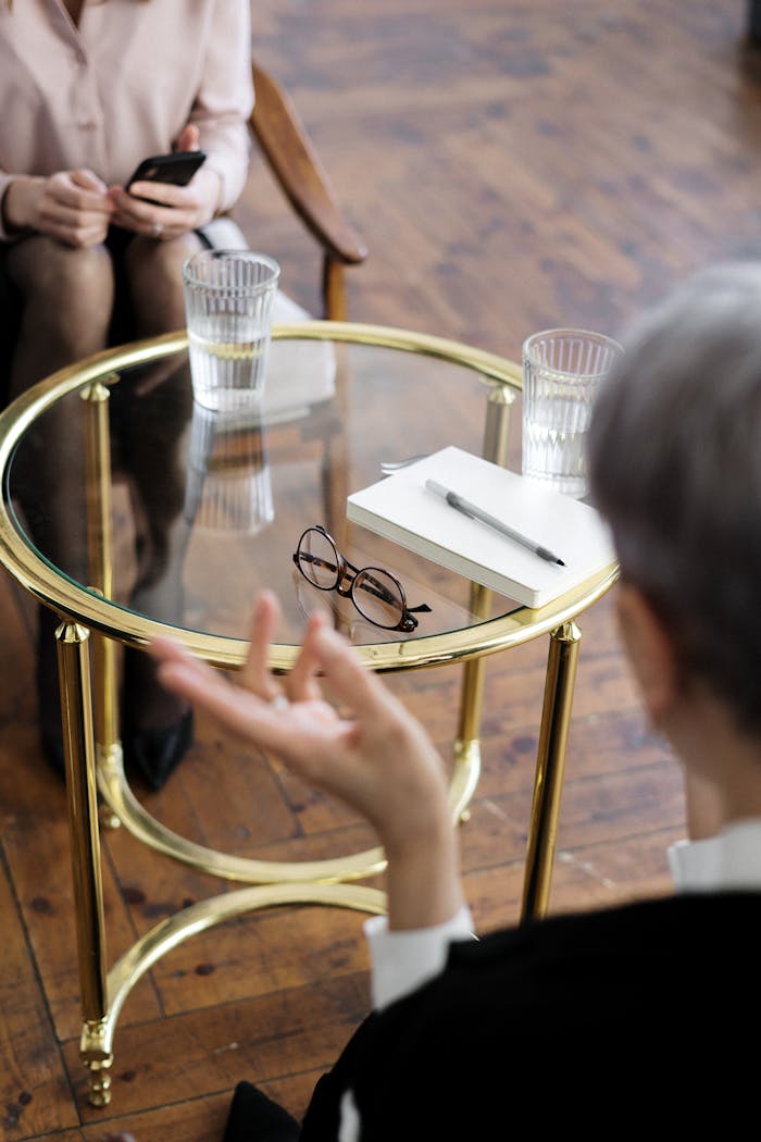 who-we-are Two adults engaged in a therapy session, seated at a glass table with notebooks and water glasses.